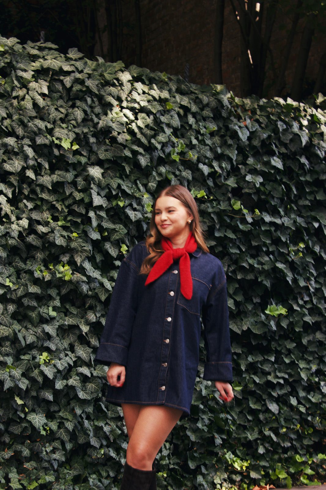 Woman wearing a navy blue dress with a red scarf standing against a wall of green ivy.