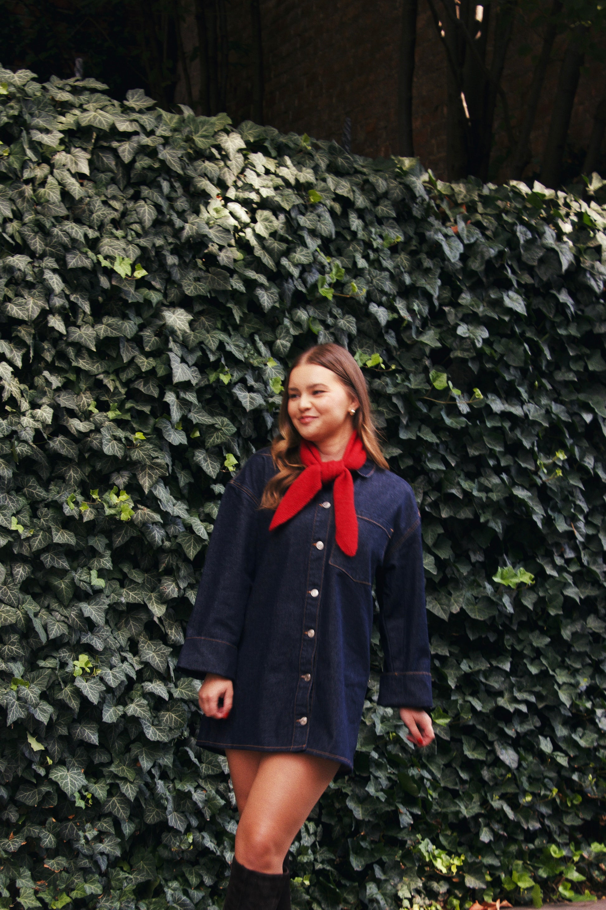 Woman wearing a navy blue dress with a red scarf standing against a wall of green ivy.
