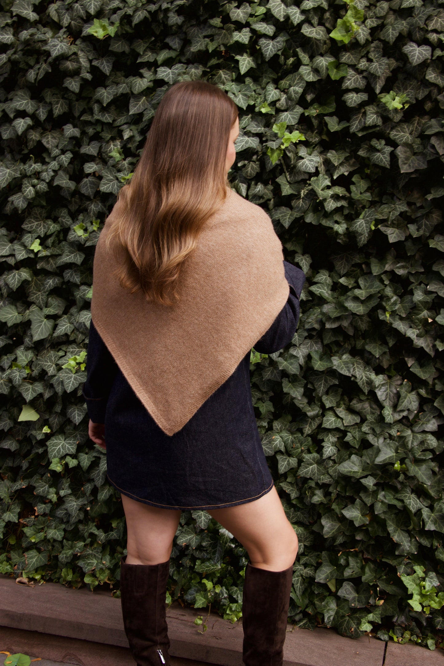 Woman wearing a beige shawl over a denim dress against a green ivy wall