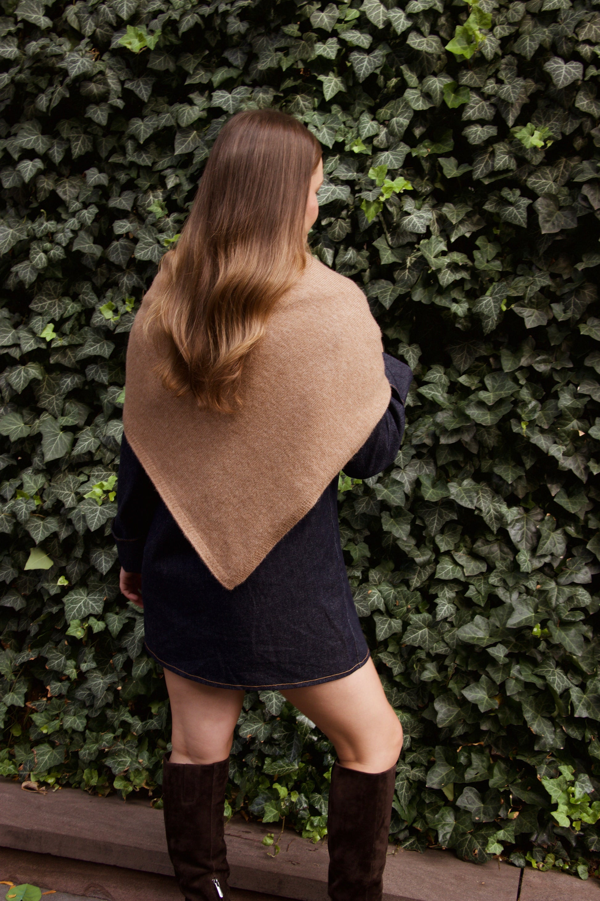 Woman wearing a beige shawl over a denim dress against a green ivy wall