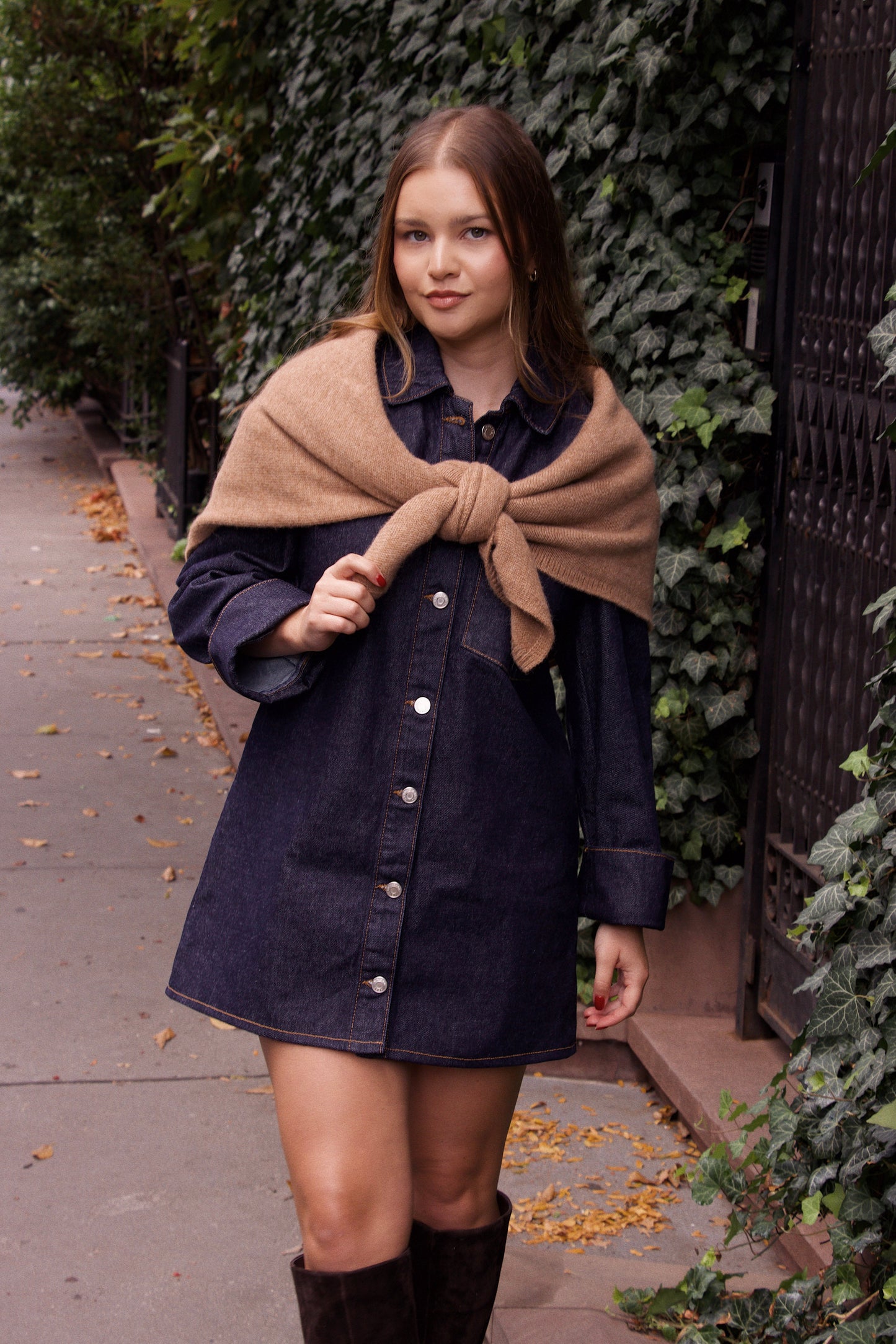 Woman wearing a denim dress with a beige scarf against a green hedge background