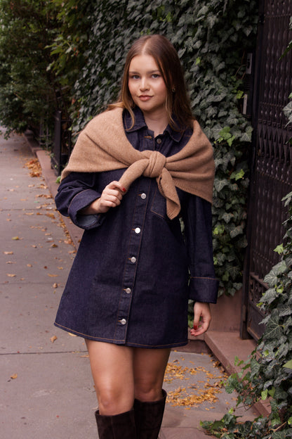 Woman wearing a denim dress with a beige scarf against a green hedge background