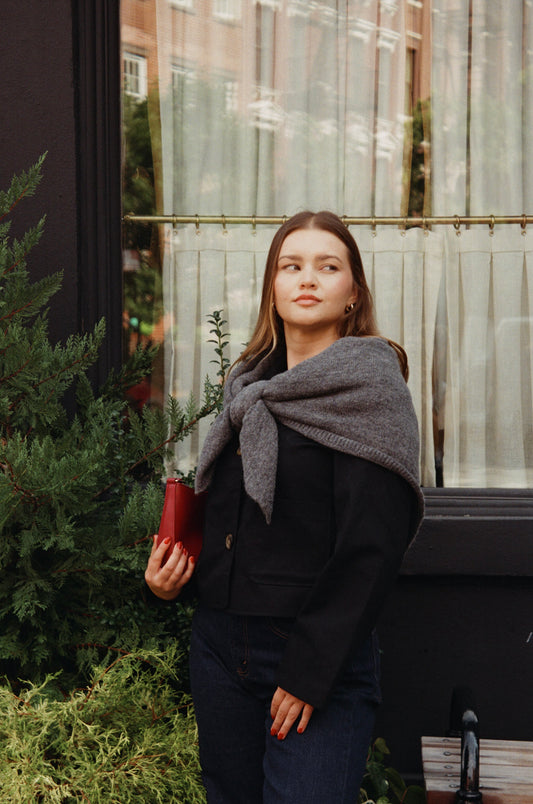 Woman wearing a gray scarf and holding a red bag in front of a window.