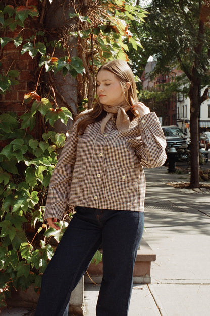 Woman wearing a plaid jacket and tan neck scarf walking outdoors on a sidewalk with trees and buildings in the background