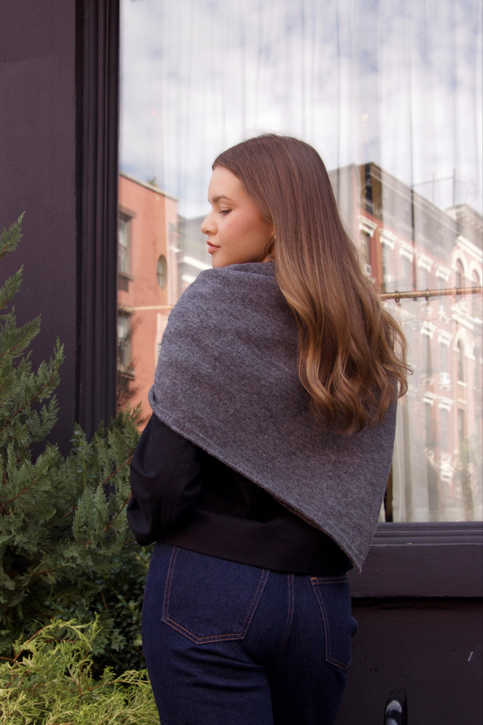 Woman wearing a gray shawl standing outdoors with greenery and a building in the background