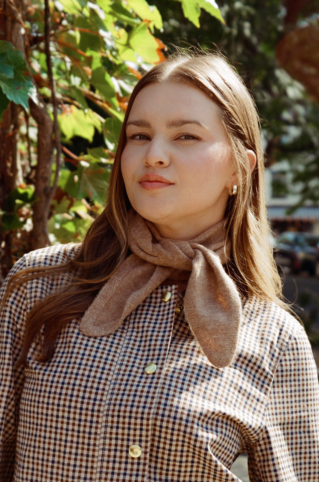Woman wearing a plaid jacket with a brown scarf, standing outdoors with greenery in the background