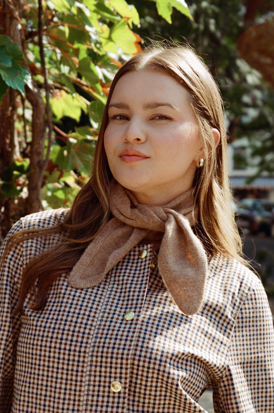 Woman wearing a plaid jacket with a brown scarf, standing outdoors with greenery in the background